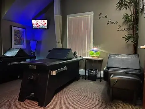 A treatment room with a padded table, a glowing round lamp on a side table, a leafy plant and the words Live, Love, Laugh on the wall
